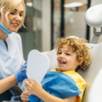 Jak przekonać dziecko do dentysty - uśmiechnięty chłopiec na fotelu dentystycznym przeglądający się w lusterku i stojąca obok dentystka