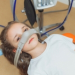 Sedacja wziewna w stomatologii - dziewczynka na fotelu stomatologicznym z aparatem do sedacji wziewnej na twarzy
