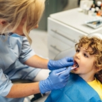 Lakierowanie zębów - chłopiec na fotelu stomatologicznym i dentystka badająca jego zęby