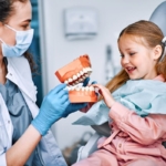 Dentysta dla dziecka - dziewczynka na fotelu stomatologicznym oglądająca model zębów trzymany przez dentystkę