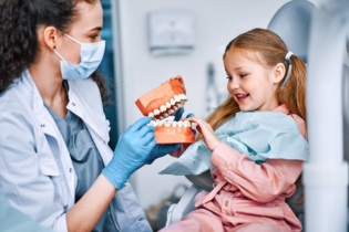 Dentysta dla dziecka - dziewczynka na fotelu stomatologicznym oglądająca model zębów trzymany przez dentystkę