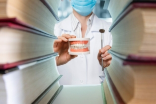 Studia stomatologiczne - człowiek w fartuchu lekarskim pomiędzy książkami trzymający lusterko dentystyczne i model szczęki