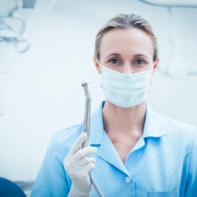 Dentysta ogólny - dentystka w gabinecie z maseczką na twarzy trzymająca narzędzie stomatologiczne